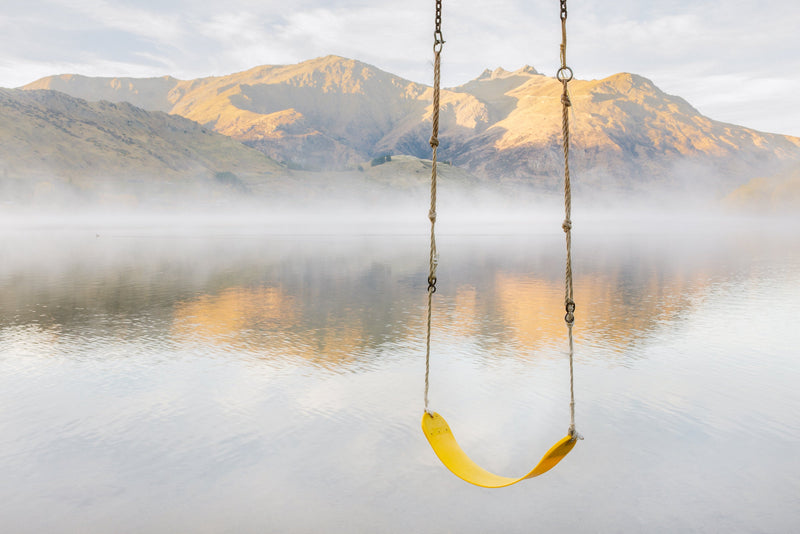 A swing rests low over the waters edge, hanging from a willow branch on the shores of Lake Hayes, Central Otago.  The road to the local skifield atop the Remarkables Mountains visible in the distance across the calm misty waters early one morning.